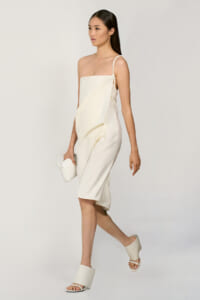 Full-body shot of a model in a cream one-shoulder draped midi dress, white clutch, and matching sandals, posing against a neutral backdrop.