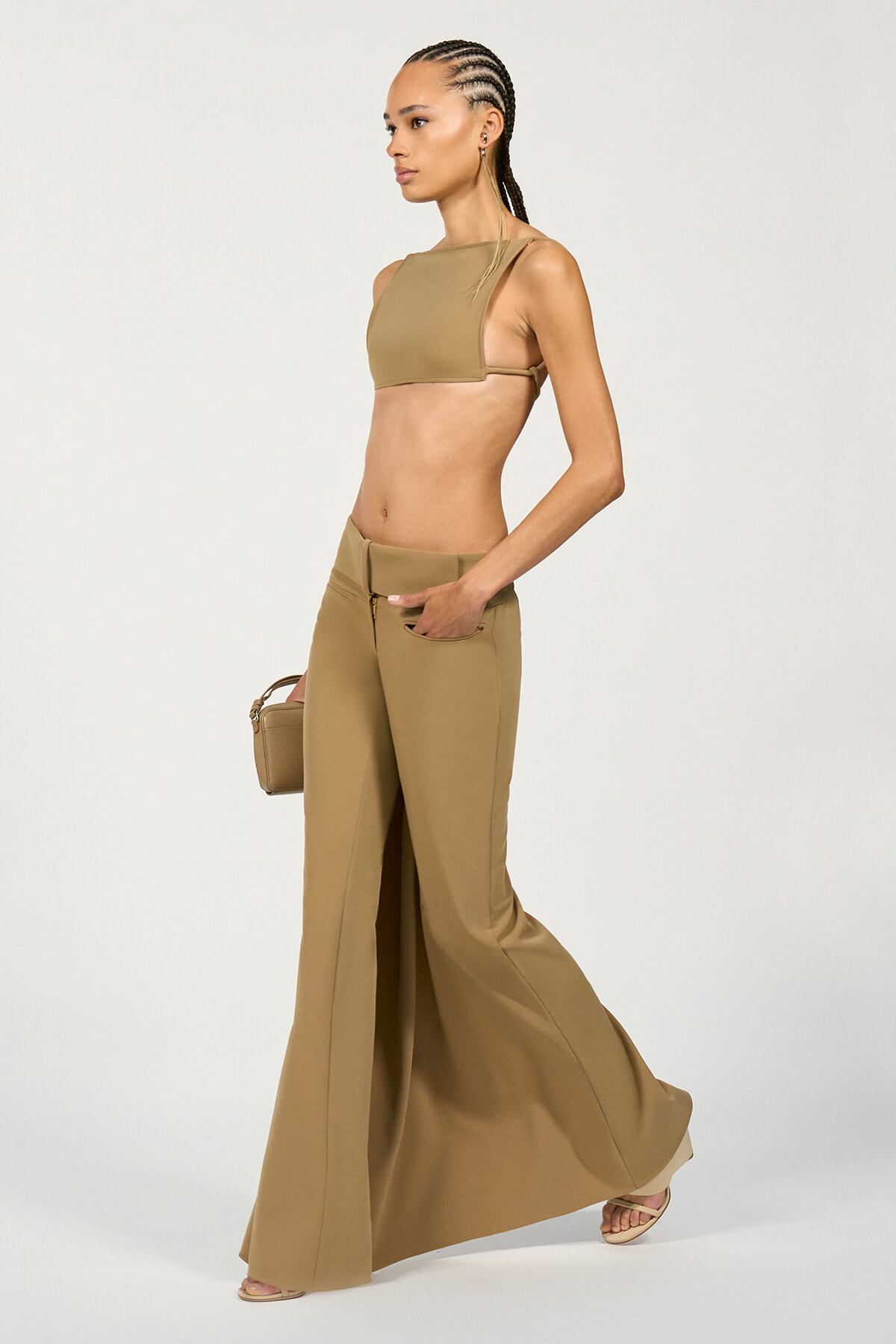 Model in a tan crop top and wide-leg pants, walking in a studio while holding a small tan handbag.