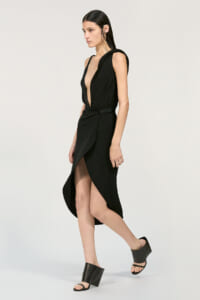 Model wearing a black sleeveless wrap dress with a deep V-neck and tied waist, paired with black block-heeled sandals.