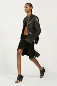 Model in black street-style outfit: leather jacket over a crop top, asymmetric fringe skirt, and chunky-heeled sandals.
