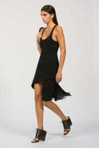 Model wearing a black sleeveless dress with an asymmetrical, high-low hem and pleated fringe at the back, walking forward in a studio setting with open-toe sandals.