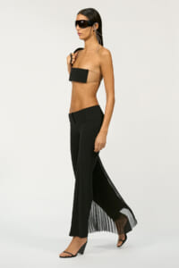 Model in a tan bandeau top with a black chest panel, black pleated-back pants, and sunglasses, walking sideways.