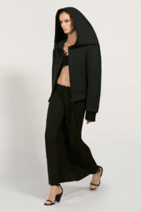 Model wearing an oversized black hooded jacket, black crop top, wide-leg pants, and strappy heels, walking forward.