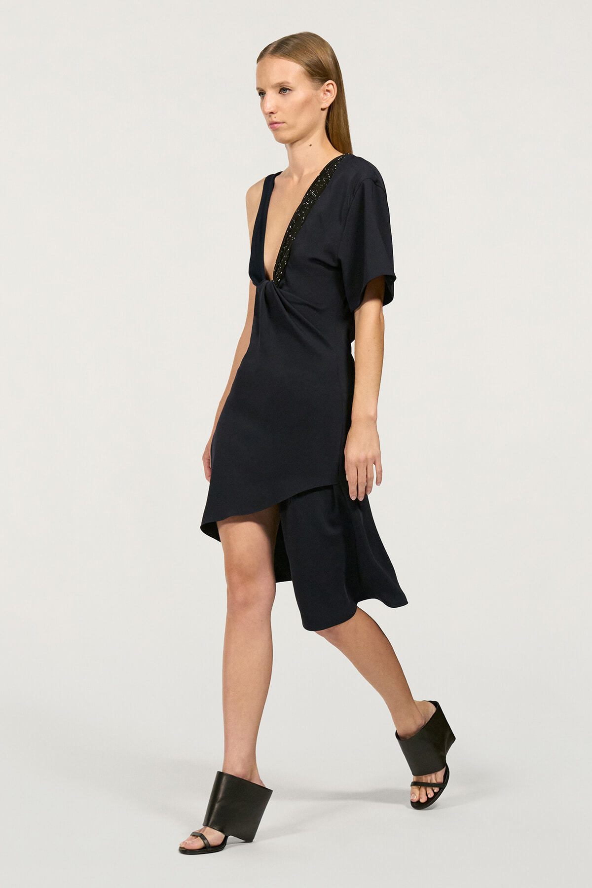 Model wearing a navy asymmetrical dress with a plunging V-neck and knot at the waist, paired with black sandals.