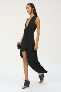Model in a black sleeveless jumpsuit/dress with deep V and asymmetric wrap skirt, carrying a small black clutch and wearing black heeled sandals with lace-up accents.