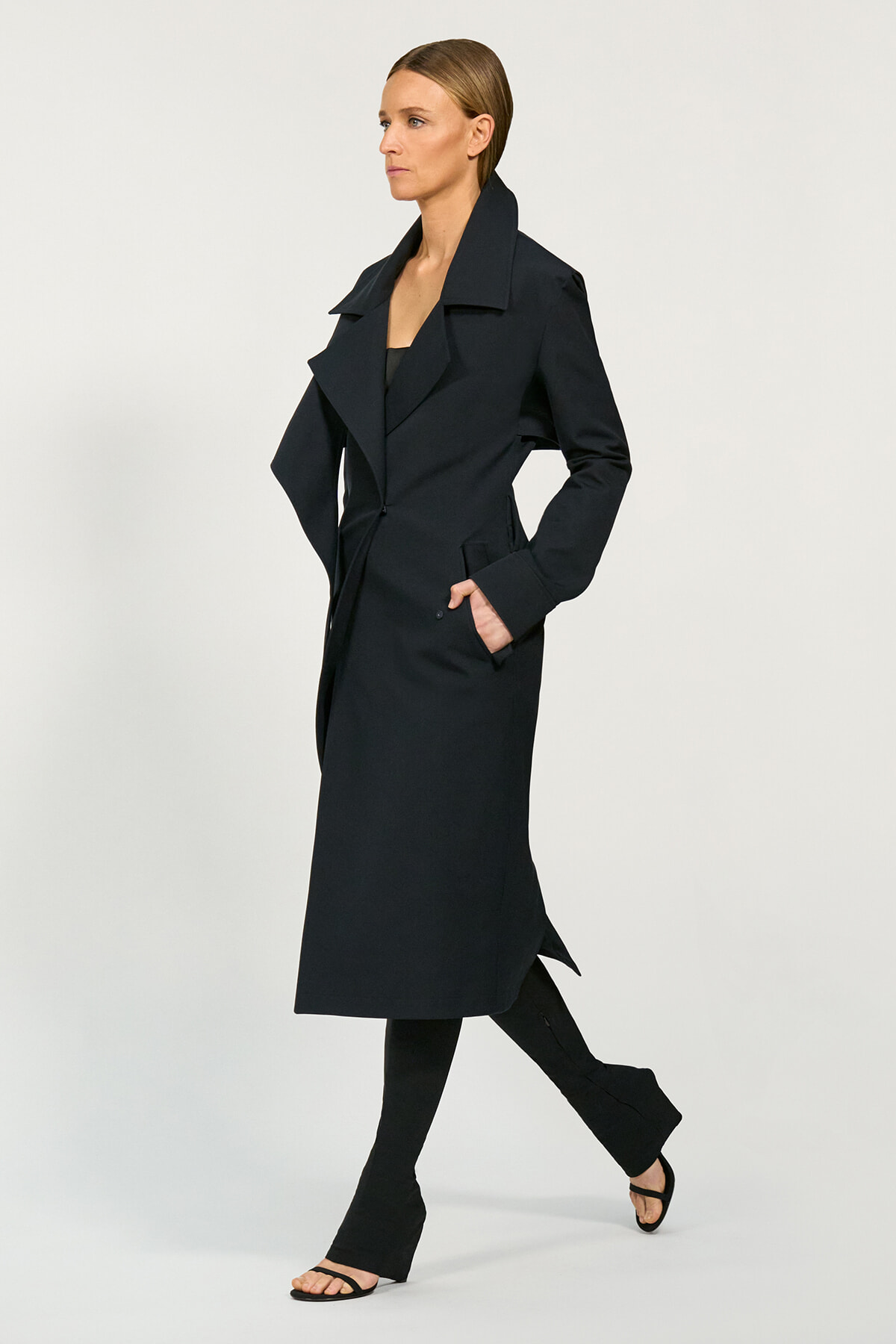 Full-length shot of a woman in a dark, knee-length trench coat with wide lapels, black outfit, and open-toe sandals, walking forward.