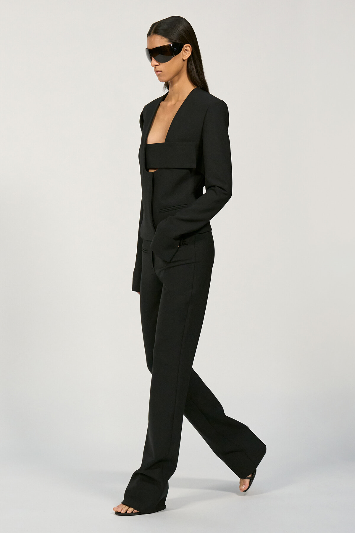 Model in a black pantsuit with a cropped blazer, wide collar, and matching wide-leg pants, wearing futuristic sunglasses and open-toe heels, walking in a studio.