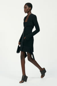Model wearing a knee‑length black coat dress with wide sleeves, a belt, and an asymmetrical hem, carrying a small black handbag.