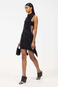 Model wearing a black beaded cutout mini dress with a side slit, carrying a small black handbag and wearing black platform sandals.