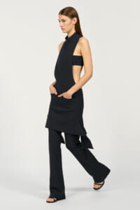 Model wearing a dark sleeveless high-neck tunic with side cutouts layered over wide-leg black pants, standing in profile.