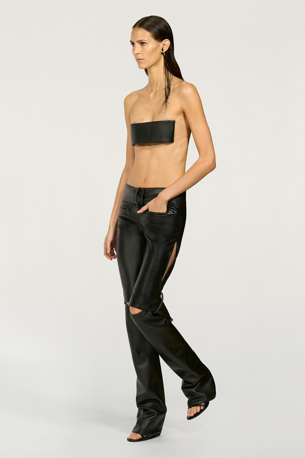 Model wearing a black bandeau-style top and matching black leather pants with side slits, posing in a studio.