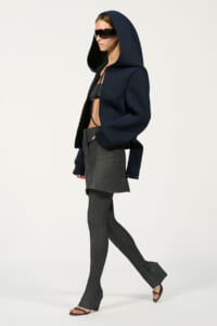 Model in a navy oversized hooded jacket, cropped top, gray layered skirt over leggings, black sandals, and large sunglasses, walking sideways.