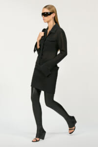 Model in an all-black outfit: long black shirt dress over black leggings, open-toe heeled sandals, and large black sunglasses, walking forward.