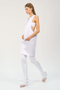 Model wearing an all-white sleeveless tunic with side cutouts over white leggings, posing in profile.