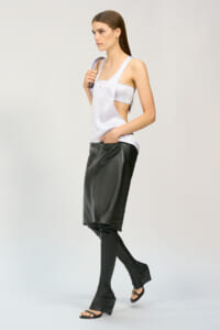 Model in a studio wearing a white sleeveless top with cutouts and a black leather knee-length skirt over black leggings, with strappy sandals.