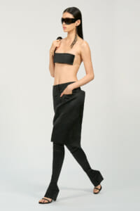 Model in a beige bandeau top with a black chest panel, paired with a black midi skirt over leggings and open-toe sandals, wearing oversized sunglasses.