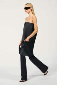 Model in a black strapless tunic over wide-leg pants, wearing black sunglasses and flat sandals, carrying a small black clutch.