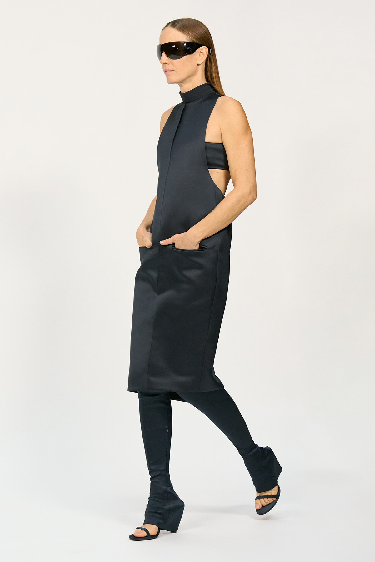 Model wearing a sleeveless dark tunic dress with high collar, side cutouts, and pockets, paired with black leggings and platform sandals, plus oversized black sunglasses.