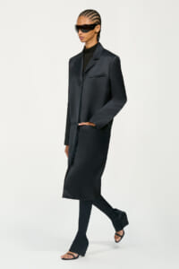 Model in a long black coat with matching trousers and open-toe sandals, wearing large futuristic sunglasses and cornrowed hair, posing mid-step.