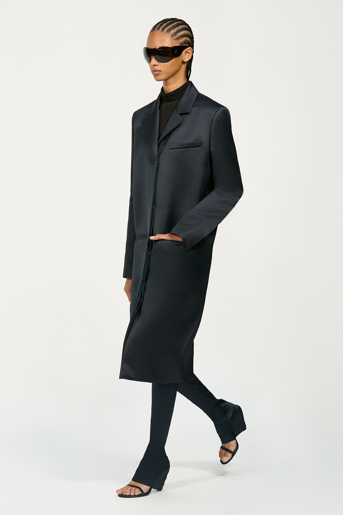 Model in a long black coat with matching trousers and open-toe sandals, wearing large futuristic sunglasses and cornrowed hair, posing mid-step.