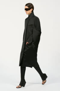 Model in a long, black, structured coat with a high collar and oversized sleeves, wearing black sunglasses and open-toe sandals against a light background.