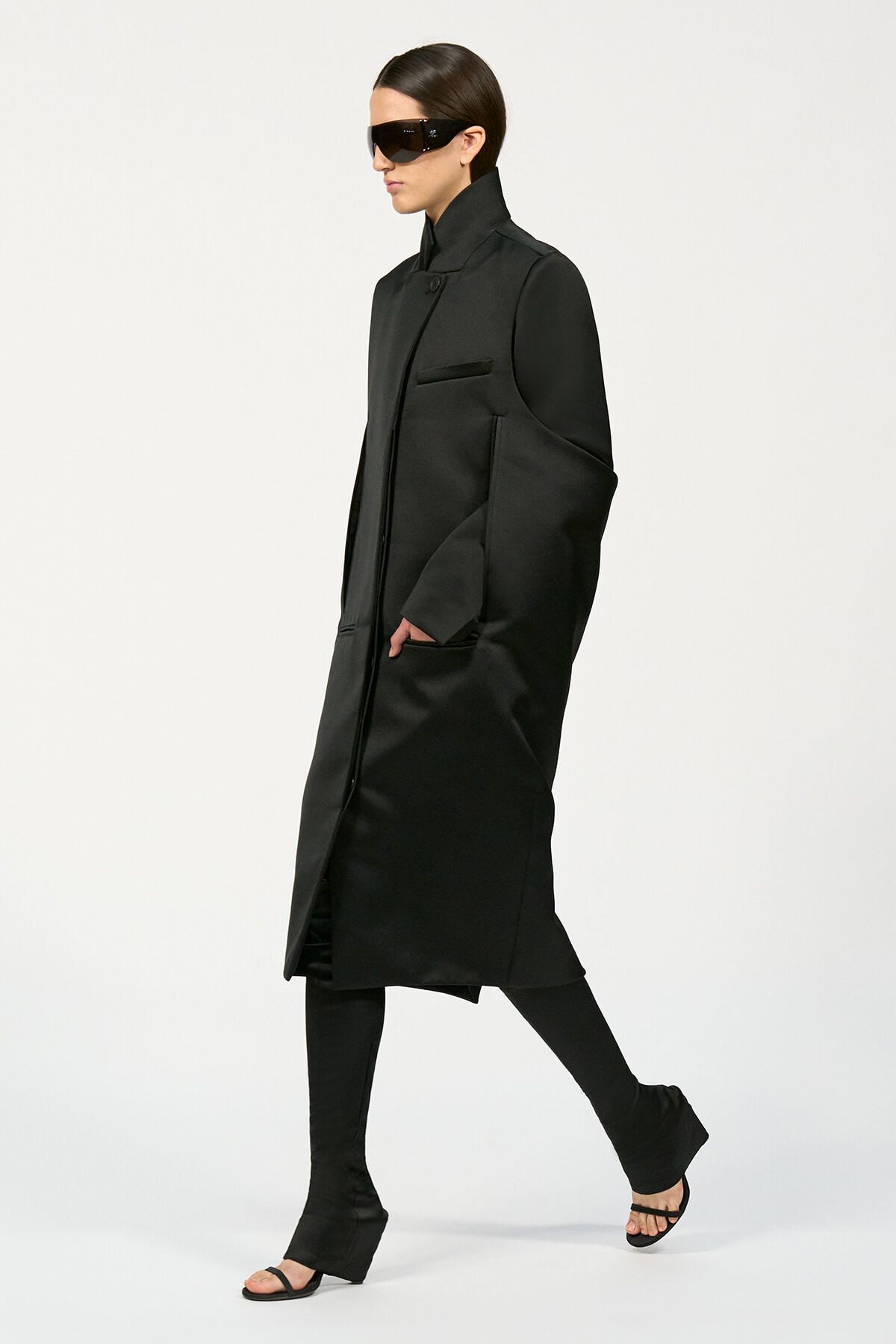 Model in a long, black, structured coat with a high collar and oversized sleeves, wearing black sunglasses and open-toe sandals against a light background.