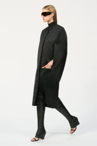 Model in a long black coat with hands in pockets, wearing oversized black sunglasses and black knee-high boots.