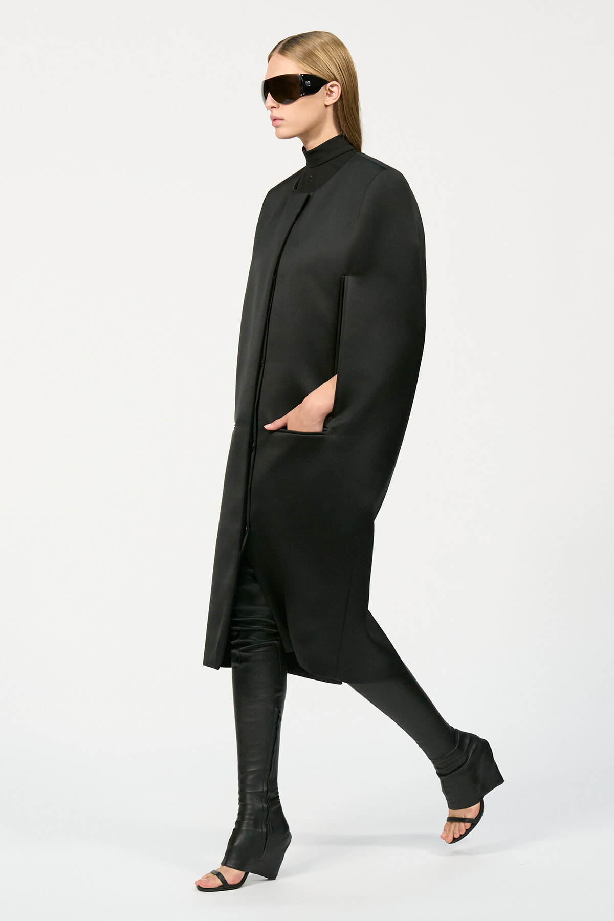 Model in a long black coat with hands in pockets, wearing oversized black sunglasses and black knee-high boots.