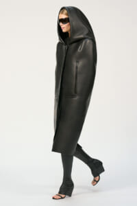 Model wearing a long black hooded leather coat with zip pockets, black leggings, and open-toe heeled sandals, in studio light.