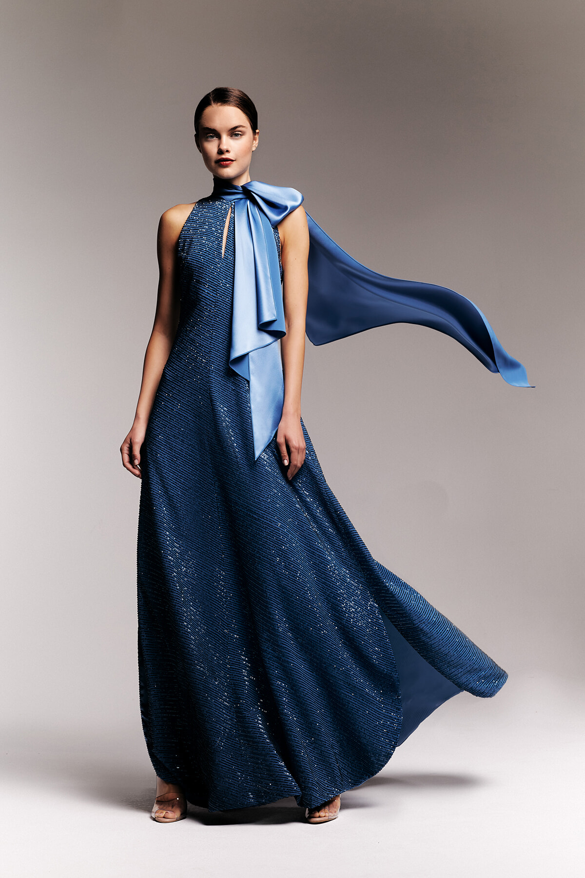 Model wearing a navy blue sequined, floor-length gown with a light blue satin scarf draped over the shoulder and flowing behind her.