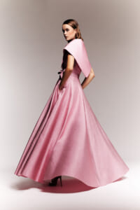 Model in a shimmering pink, floor-length ball gown with a cape-like shoulder and belt, posing in profile for a fashion shoot.