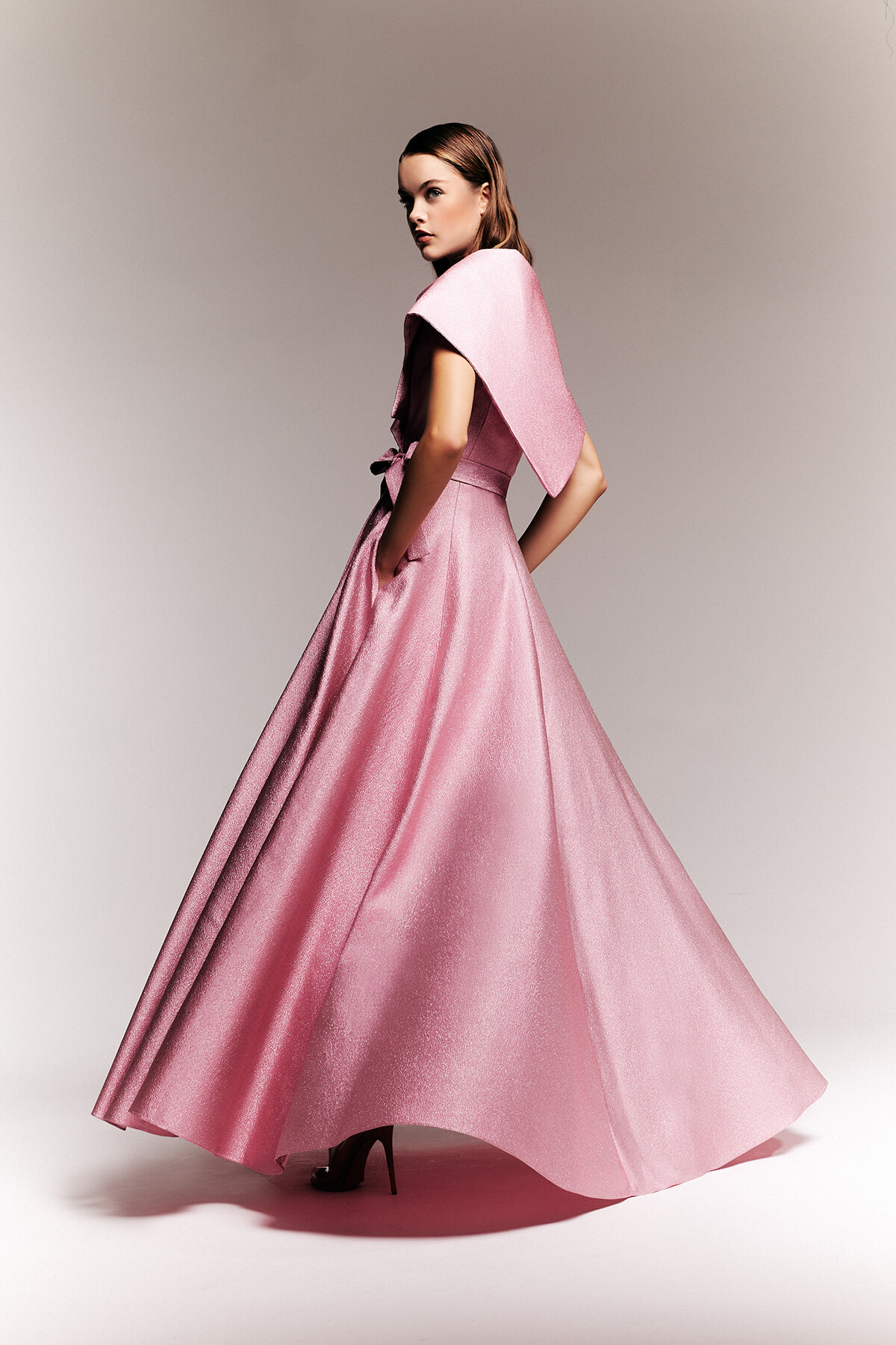 Model in a shimmering pink, floor-length ball gown with a cape-like shoulder and belt, posing in profile for a fashion shoot.