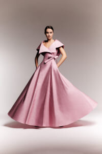 Model wearing a pink, floor-length gown with a dramatic wide collar and bow at the waist, spinning to showcase the full skirt.