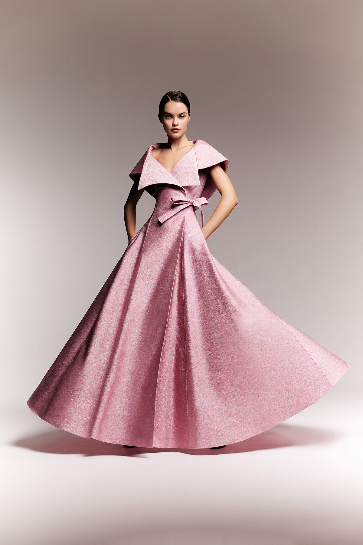 Model wearing a pink, floor-length gown with a dramatic wide collar and bow at the waist, spinning to showcase the full skirt.