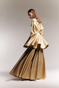 Side-profile of a model in a shimmering gold, floor-length gown with a voluminous, flowing skirt and cape-like bodice in a studio setting.