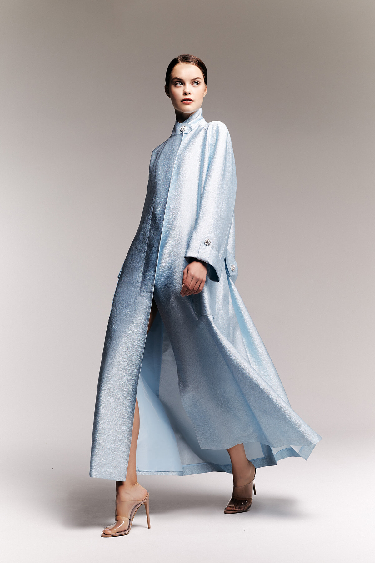 Model in a pale blue metallic long coat with high collar, flowing to the floor, paired with nude heeled sandals; fashion shoot.
