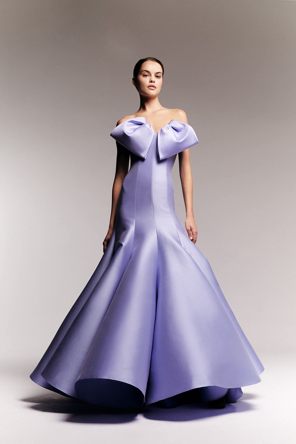 Woman wearing a lavender off-shoulder ball gown with a large bow at the neckline and a full, satin skirt.
