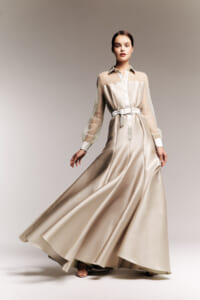 Model wearing a long champagne satin dress with sheer dotted sleeves and a wide belt, posing mid-stride against a neutral backdrop.