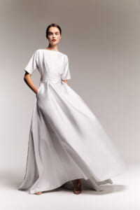 Model wearing a white, floor-length gown with short sleeves and a wide waist sash, posing against a neutral backdrop.