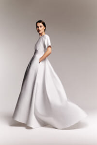 Model wearing a white, high-fashion gown with short sleeves and a full, flowing skirt, posing mid-walk against a neutral backdrop.