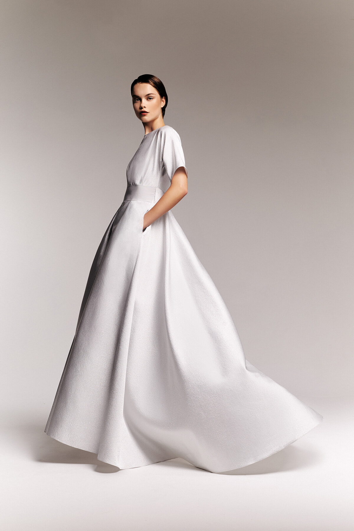 Model wearing a white, high-fashion gown with short sleeves and a full, flowing skirt, posing mid-walk against a neutral backdrop.