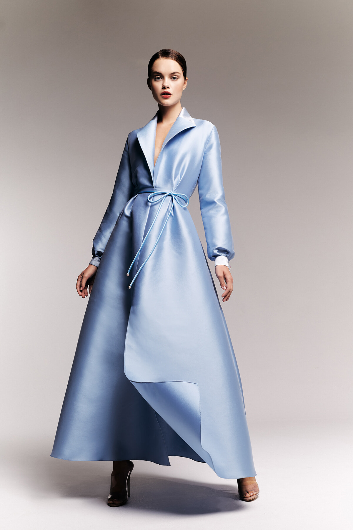 Model wearing a light-blue satin wrap coat with a waist tie, floor-length and styled against a neutral studio backdrop.