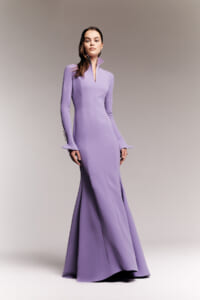 Model wearing a lavender, floor-length mermaid gown with a high collar and flared cuffs, posed against a neutral gradient backdrop.