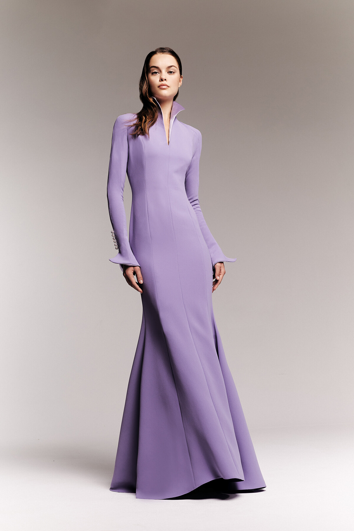 Model wearing a lavender, floor-length mermaid gown with a high collar and flared cuffs, posed against a neutral gradient backdrop.