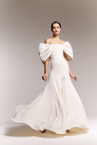 Model wearing an off-the-shoulder white gown with dramatic oversized puff sleeves, walking on a neutral studio backdrop, the dress flowing elegantly to the floor.