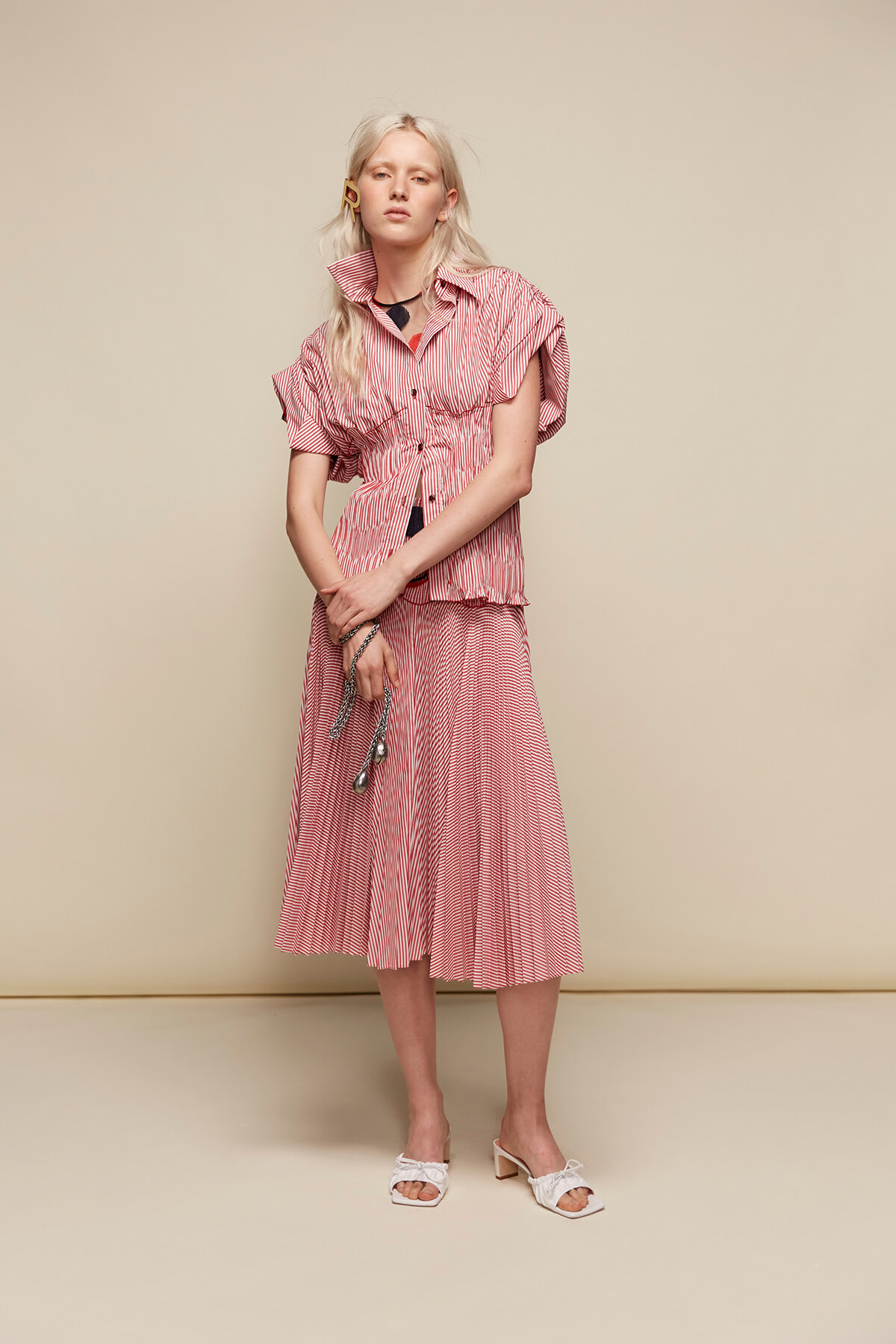 Woman with long blonde hair wearing a pink-red striped dress, standing against a beige backdrop, hands clasped, white heeled sandals.