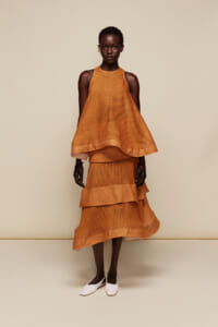 Female model wearing a bright orange sleeveless halter-neck mesh top with a layered midi skirt, standing against a beige backdrop.