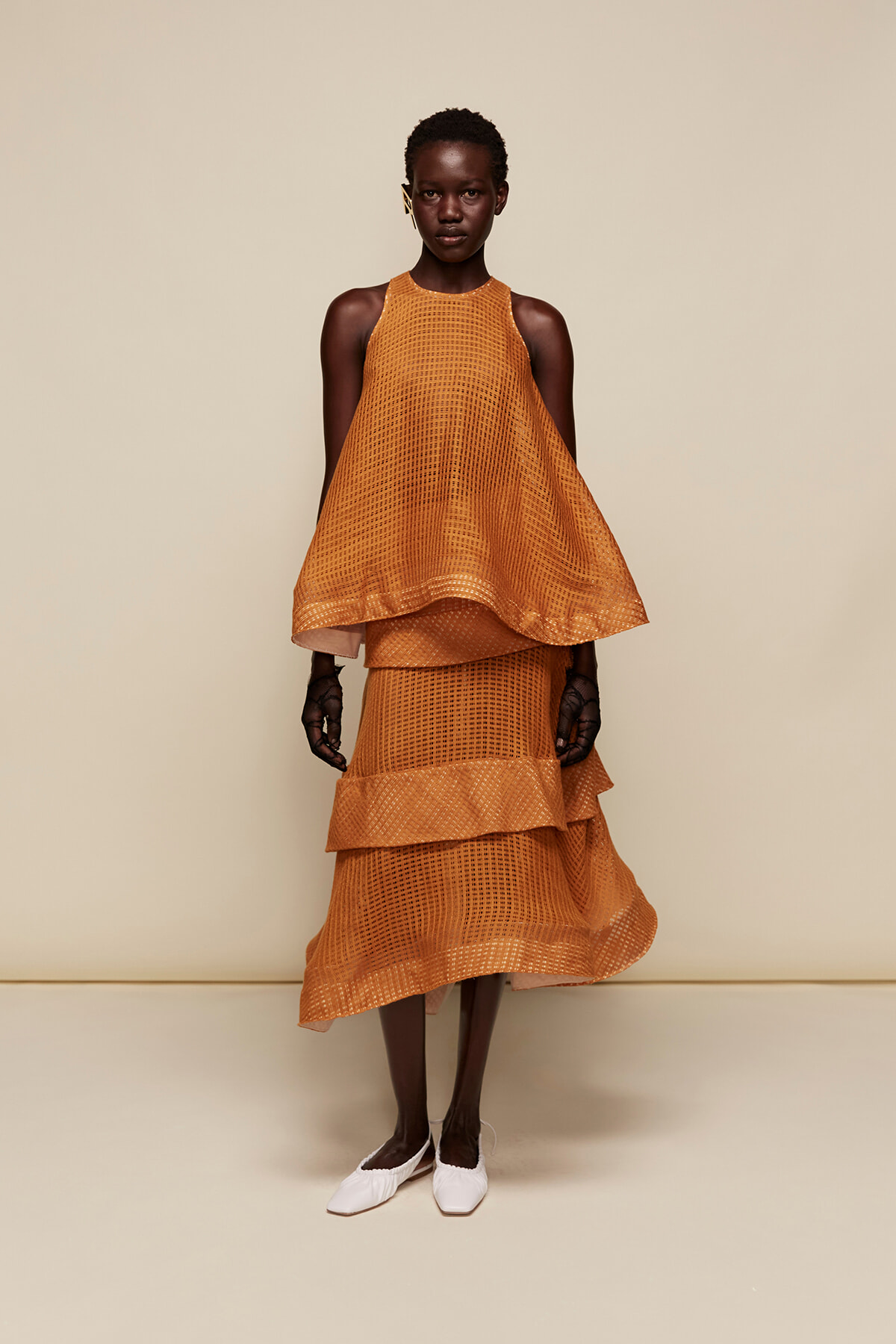 Female model wearing a bright orange sleeveless halter-neck mesh top with a layered midi skirt, standing against a beige backdrop.