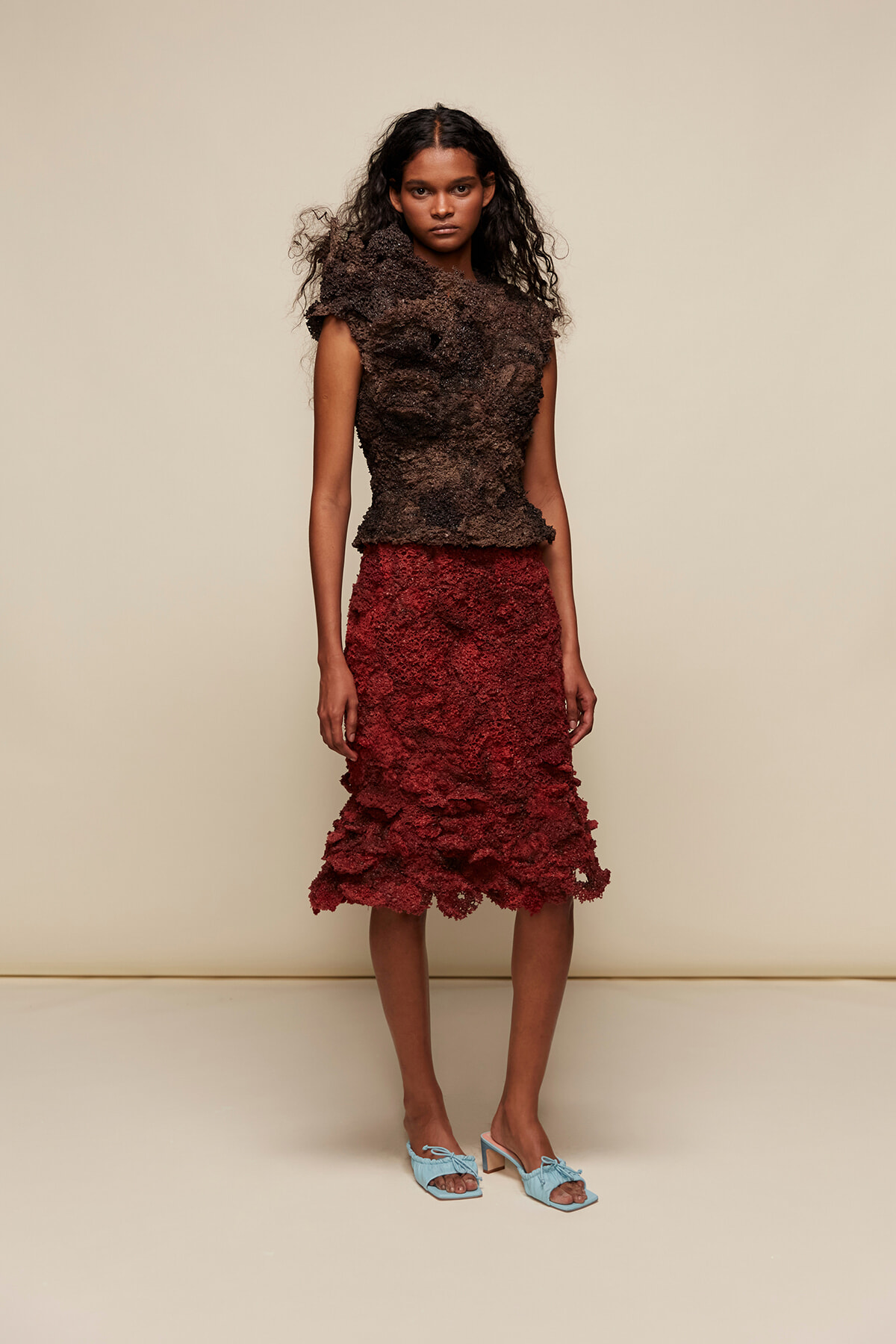 Female model in a dark, sculptural textured top and a red knee-length textured skirt, wearing light blue heeled sandals, standing against a beige studio backdrop.