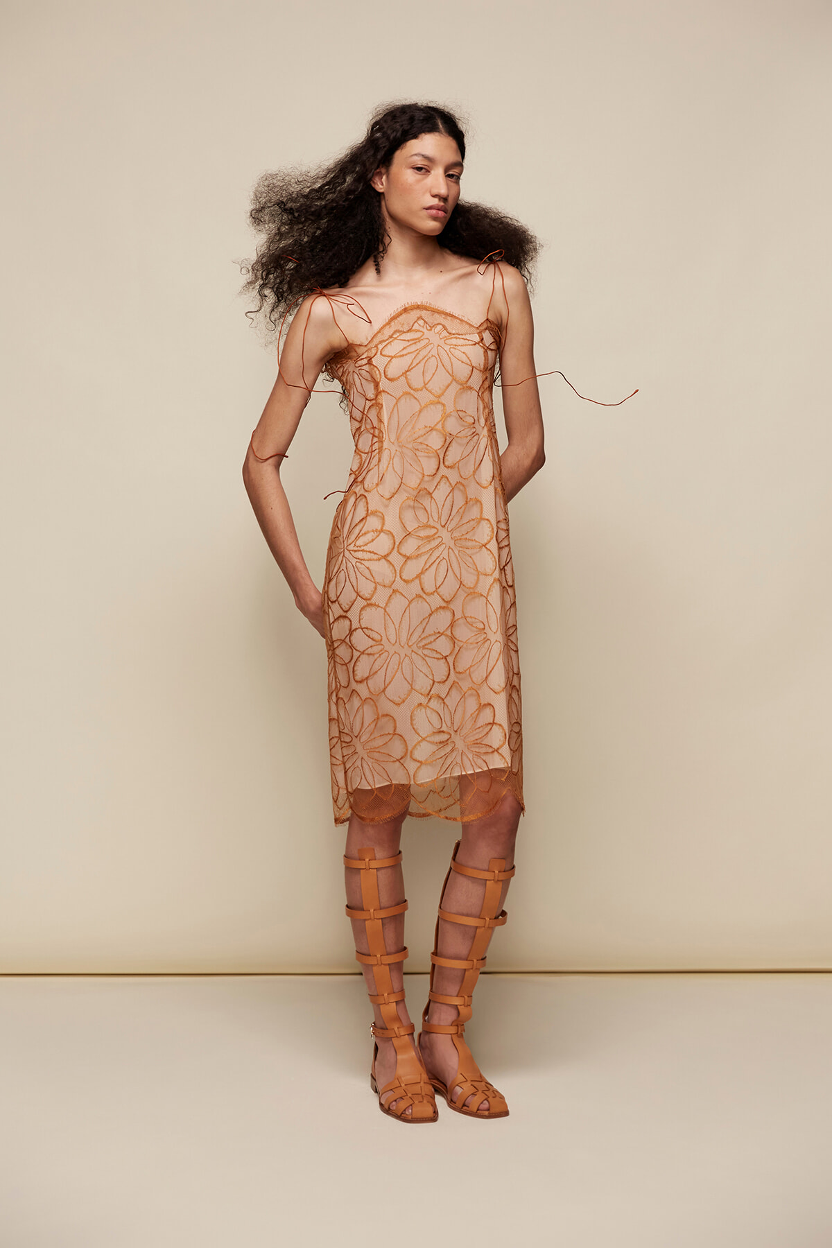 Model wearing a sheer peach dress with orange floral embroidery and matching tan gladiator sandals.